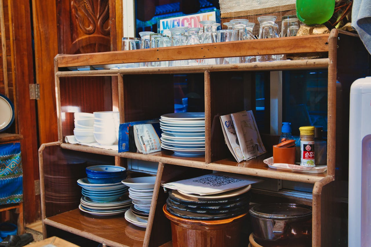 about-us A wooden kitchen shelf stocked with assorted dishes, glasses, and condiments. Cozy vintage style.